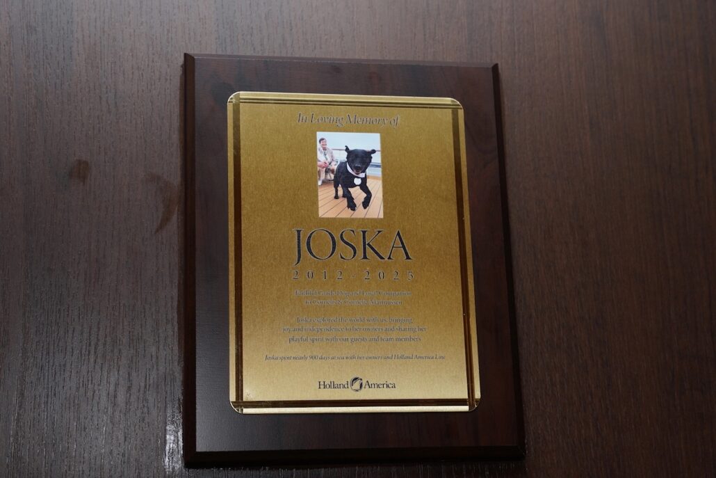A wooden plaque with a gold plate featuring a service dog’s photo at the top and the inscription “In Loving Memory of JOSKA 2012-2023,” dedication text, and the Holland America logo at the bottom, honoring loyal cruise guests.