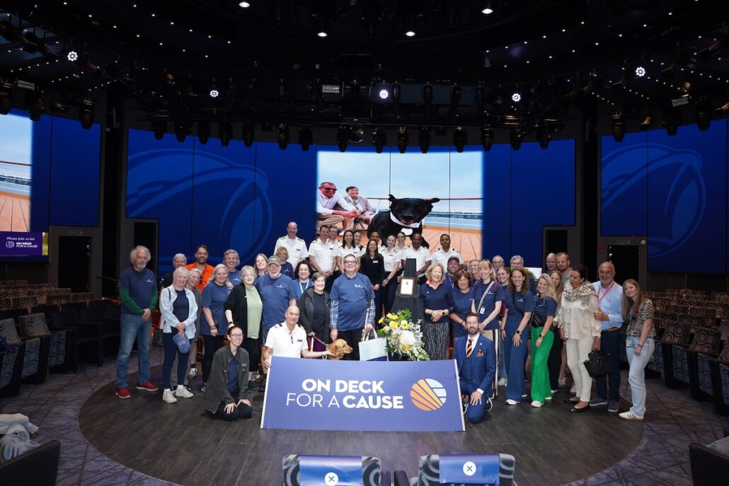 A large group of Holland America cruise guests poses on stage with a sign reading "On Deck For A Cause"; behind them, a photo featuring people with a service dog is displayed in a modern auditorium.