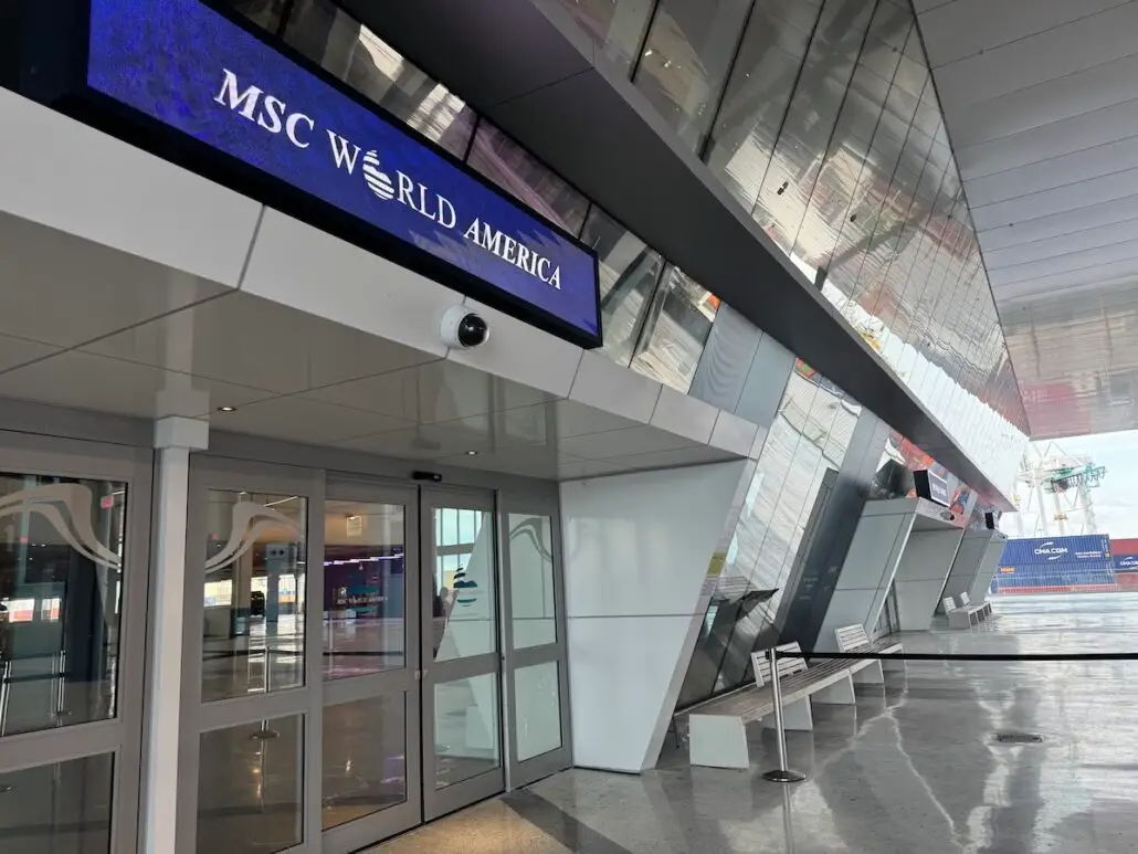 Entrance to a modern building with glass doors and a digital sign reading "MSC World America." Inside, reflective surfaces, benches, and large windows reveal a docked ship—perfect for cruise ship comparison with the Royal Caribbean Oasis Class.