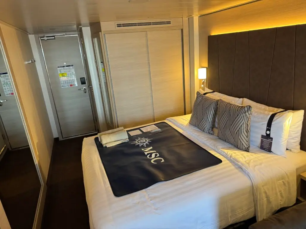 A modern cruise ship cabin aboard MSC World America features a large neatly made bed with multiple pillows, a black MSC-branded runner, informational papers, a towel, and key card. Light wood furnishings and a mirrored closet complete the look.
