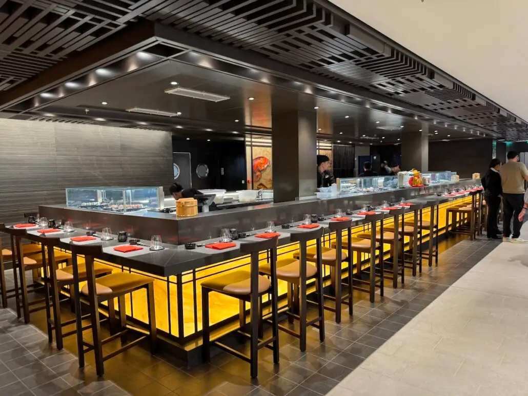 A modern sushi restaurant, reminiscent of Royal Caribbean Oasis Class dining, features a long illuminated bar with high wooden chairs, red-napkin place settings, and several chefs preparing food behind a glass counter. Diners are visible at the far end.