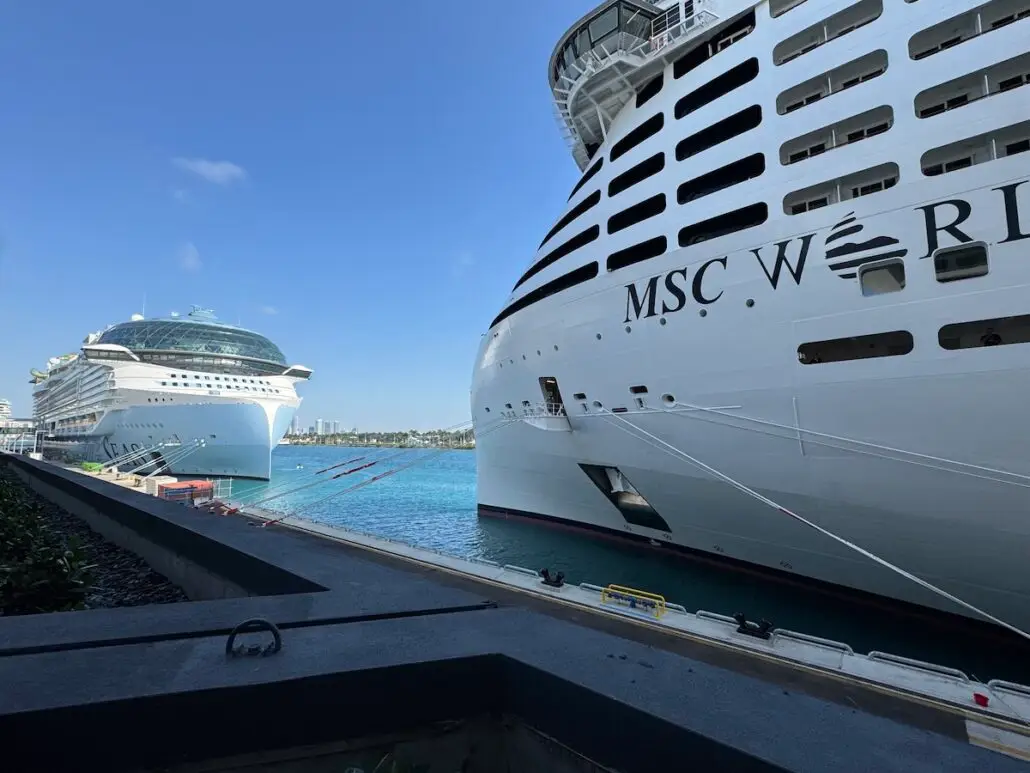 Two large cruise ships are docked side by side at a port, with "MSC W" visible on the hull of the MSC World America. The clear blue sky and calm harbor set the scene for a striking cruise comparison moment.