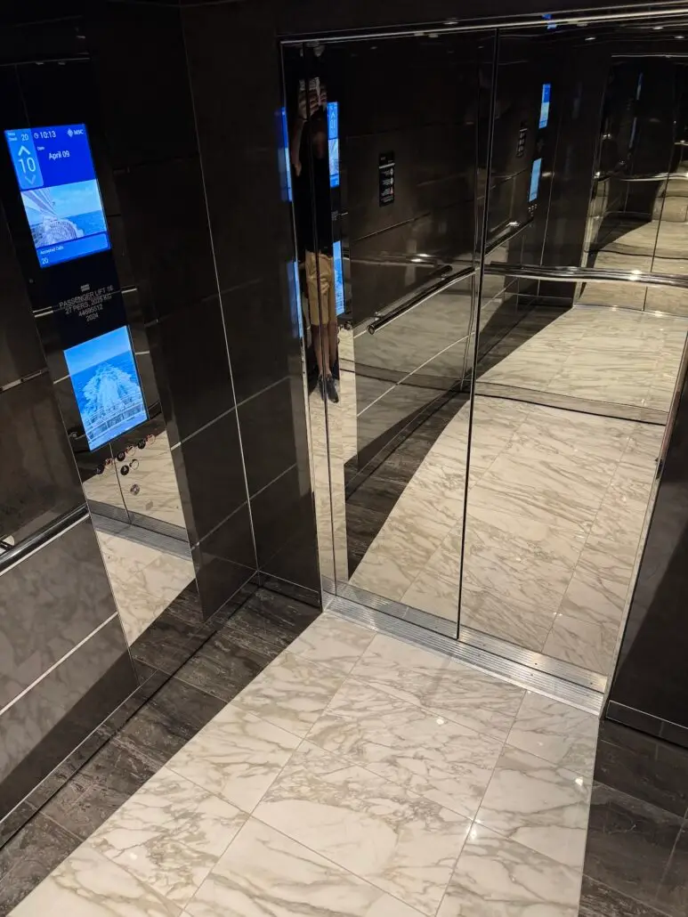 A modern elevator interior with marble tile flooring, dark reflective walls, and a digital display—like those on MSC World America—shows the floor number and a mountain image, with a partial reflection visible in the mirrored door.