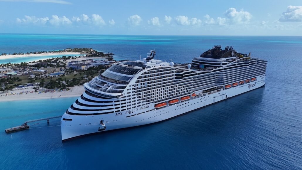 A large white Royal Caribbean Oasis Class cruise ship is docked at a pier near a tropical island with sandy beaches, palm trees, and clear blue water under a sunny sky with scattered clouds.
