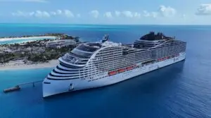 A large white Royal Caribbean Oasis Class cruise ship is docked at a pier near a tropical island with sandy beaches, palm trees, and clear blue water under a sunny sky with scattered clouds.