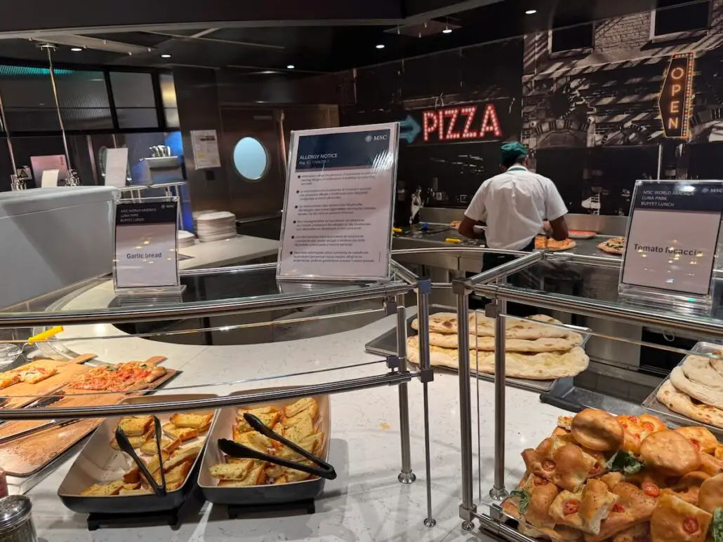A buffet counter on MSC World America displays various types of pizza, garlic bread, and calzones. A chef works behind the counter under a large "PIZZA" sign, with allergen notices posted nearby for guests' safety.