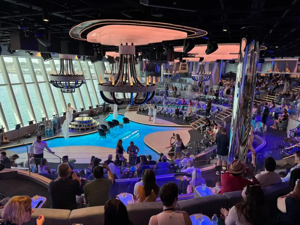 A large, modern cruise ship lounge with high ceilings, large windows facing the water, and multiple tiers of seating—reminiscent of Royal Caribbean Oasis Class venues. People gather around a central stage under impressive circular chandeliers.