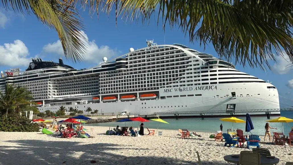 A large white cruise ship, MSC World America, is docked near a sandy beach with palm trees, colorful umbrellas, and lounge chairs—offering a vibrant scene reminiscent of the Royal Caribbean Oasis Class experience.
