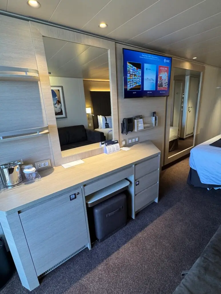 A modern hotel room with light wood furnishings, a large mirror, wall-mounted TV showing cruise comparison content, mini fridge, desk area, partially visible bed, and doorway leading to an adjoining room.