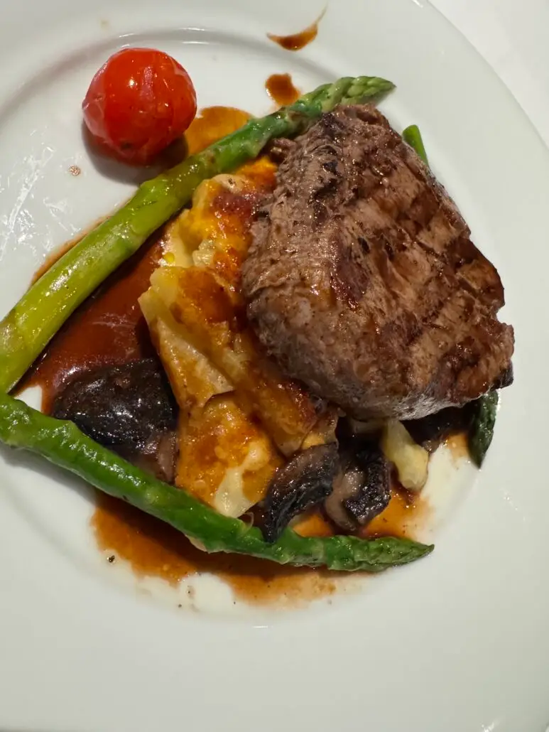 A grilled steak served on top of sliced potatoes and mushrooms, garnished with two asparagus spears and a roasted cherry tomato, with brown sauce on a white plate—perfect for savoring aboard MSC World America.