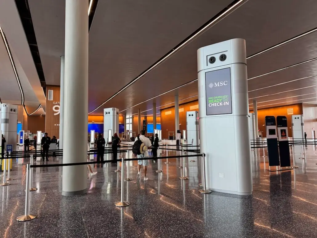 People stand in line at a modern cruise terminal with high ceilings, bright lighting, and digital check-in kiosks. Large columns display "MSC" and "Ready to Check-In," setting the stage for an MSC World America vs. Royal Caribbean Oasis Class cruise comparison.