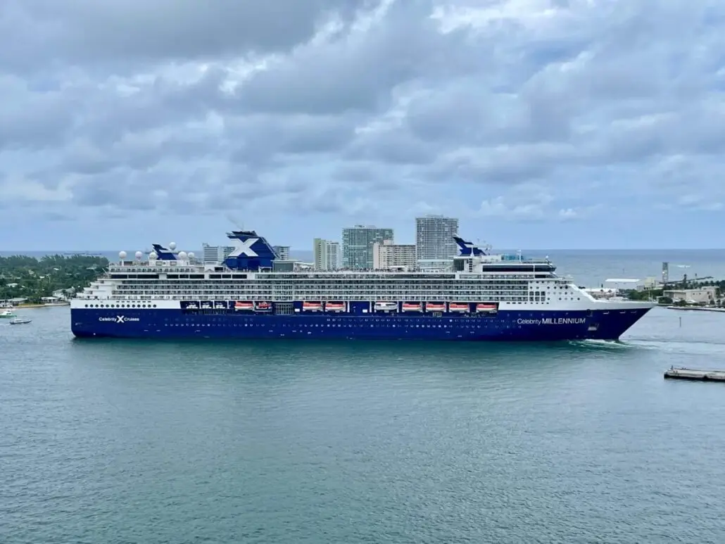 A large blue and white Celebrity Cruises ship labeled "Celebrity Millennium" sails near a city shoreline with tall buildings under a cloudy sky. The calm ocean and a small pier are visible in the foreground.