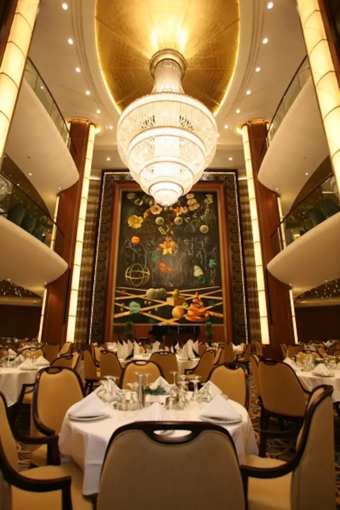 Elegant dining room with neatly set round tables, tan chairs, a large crystal chandelier, and a decorative wall mural—reminiscent of the grandeur found on Royal Caribbean Oasis Class ships. Two upper balconies offer splendid views of the space.