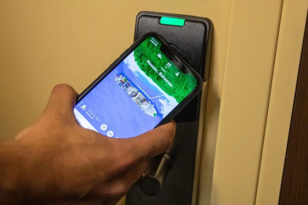 A person holds a smartphone displaying a digital room key app against an electronic door lock with a green indicator light, similar to the seamless cabin entry experience on MSC World America and Royal Caribbean’s Oasis Class ships.