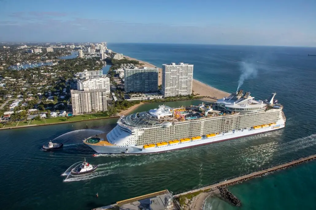 A large MSC World America cruise ship is escorted by two tugboats as it departs from a harbor near a city with high-rise buildings and a beach coastline under a clear sky.