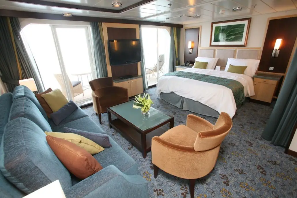 A modern hotel room inspired by MSC World America, featuring a king-size bed, seating area with blue sofa and tan chairs, glass coffee table with flowers, wall-mounted TV, and a balcony with outdoor chairs visible through glass doors.