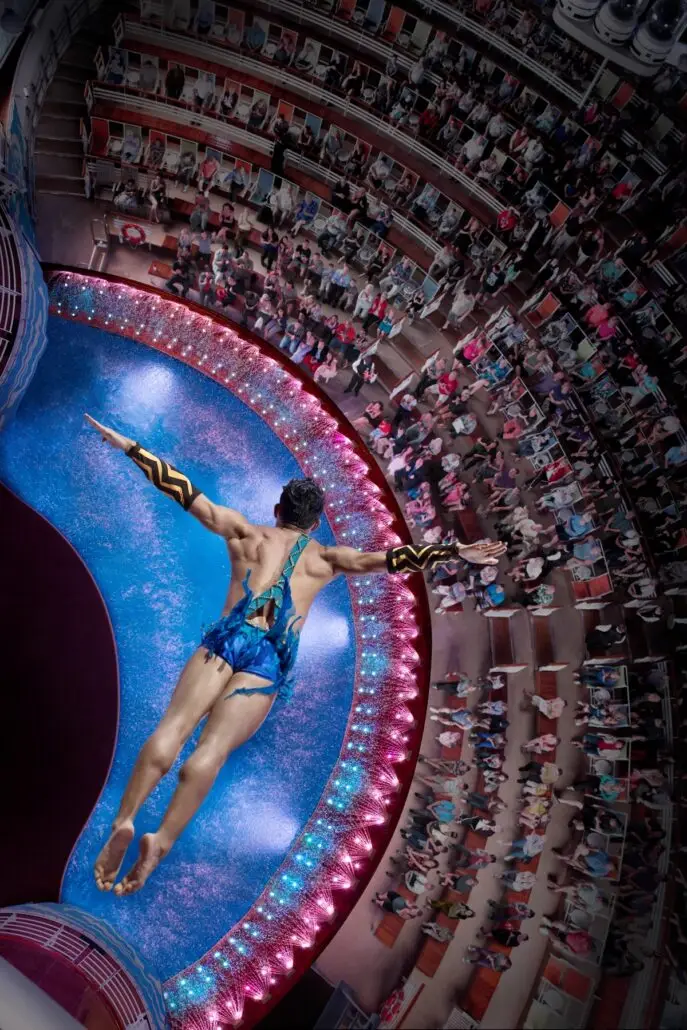 A male diver in a blue costume leaps into a circular pool from a great height, arms outstretched, as spectators fill the curved seating—reminiscent of the thrilling AquaTheater shows on Royal Caribbean Oasis Class cruise ships.