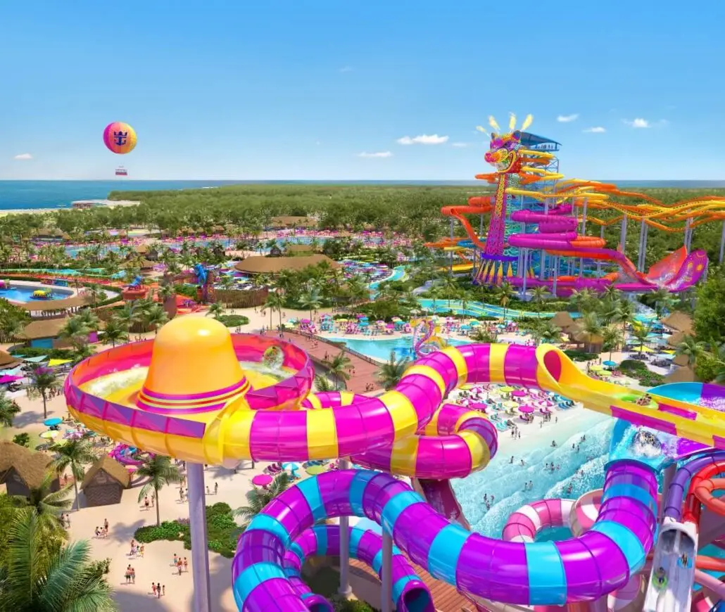 Aerial view of a colorful water park with winding pink, yellow, and purple water slides, pools, palm trees, lounge areas, and a hot air balloon in the background near the ocean.