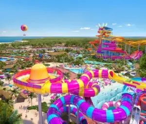 Aerial view of a colorful water park with winding pink, yellow, and purple water slides, pools, palm trees, lounge areas, and a hot air balloon in the background near the ocean.