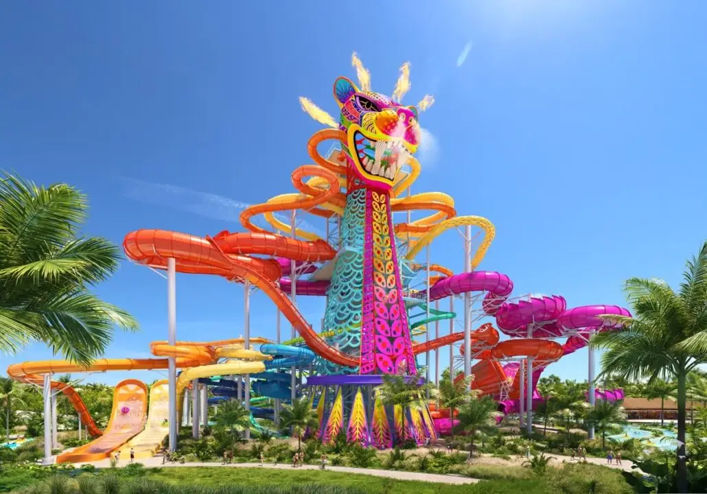 A large, colorful water slide structure shaped like a dragon, with several winding slides in orange, pink, and yellow, stands amid palm trees under a clear blue sky at a water park.