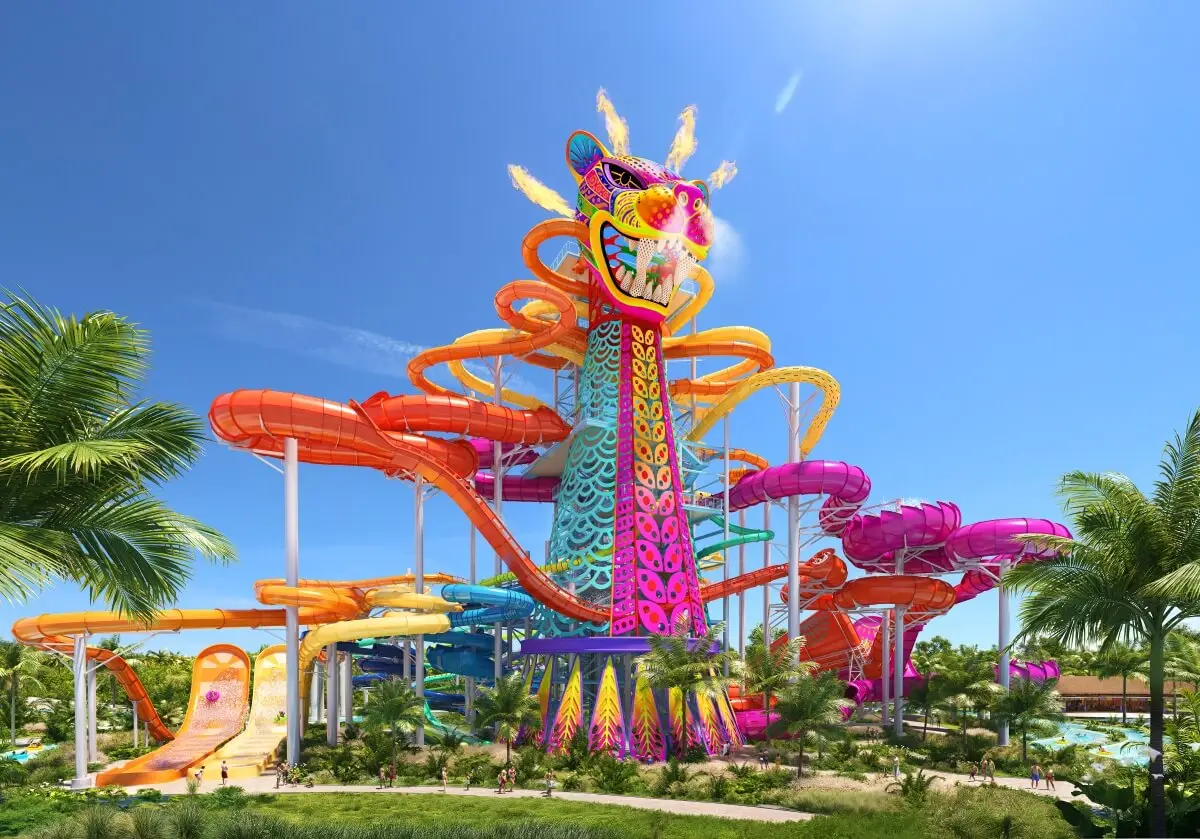 A large, colorful water slide structure shaped like a dragon, with several winding slides in orange, pink, and yellow, stands amid palm trees under a clear blue sky at a water park.