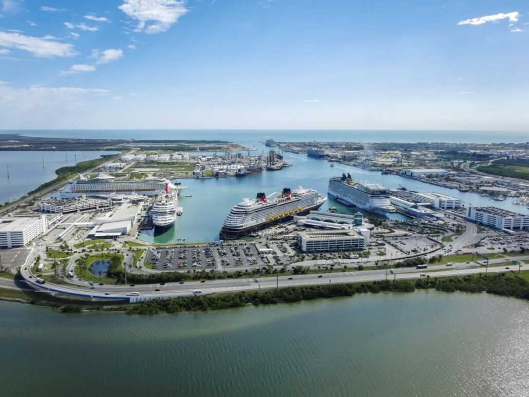 Aerial view of Port Canaveral with several cruise ships docked at upgraded cruise terminal facilities, parking lots filled with cars, and multiple buildings nearby, bordered by water and green landscaped areas under a clear sky.