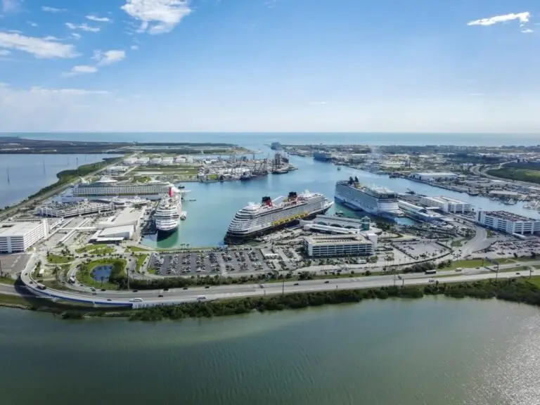 Aerial view of Port Canaveral with several cruise ships docked at upgraded cruise terminal facilities, parking lots filled with cars, and multiple buildings nearby, bordered by water and green landscaped areas under a clear sky.