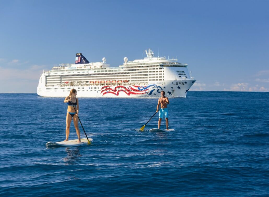 Two people paddleboard on calm blue water with the Norwegian Epic cruise ship in the background. The sky is clear and sunny, hinting at relaxing days ahead exploring new suites and exclusive adults-only spaces.