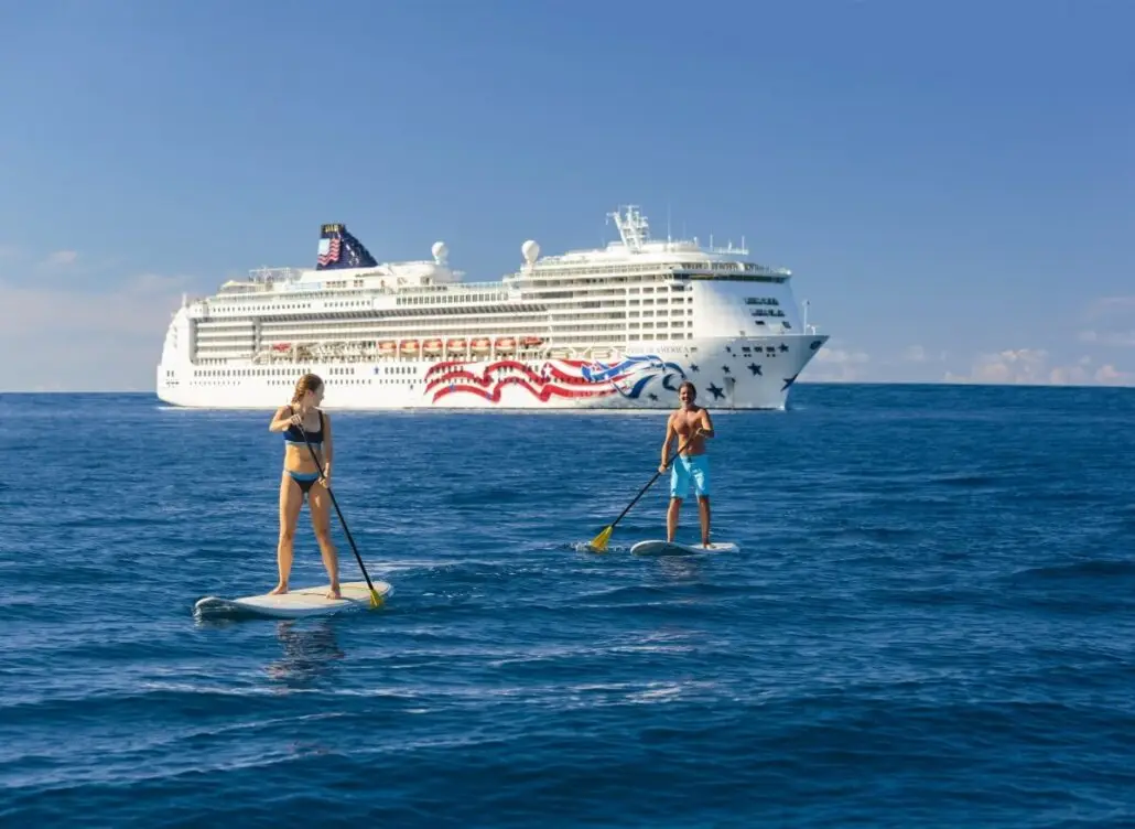 Two people paddleboard on calm blue water with the Norwegian Epic cruise ship in the background. The sky is clear and sunny, hinting at relaxing days ahead exploring new suites and exclusive adults-only spaces.