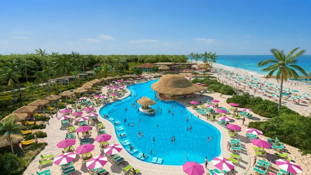A large resort pool with people swimming, surrounded by green lounge chairs and pink umbrellas. There are thatched-roof huts, palm trees, and a sandy beach with turquoise ocean in the background under a clear blue sky.