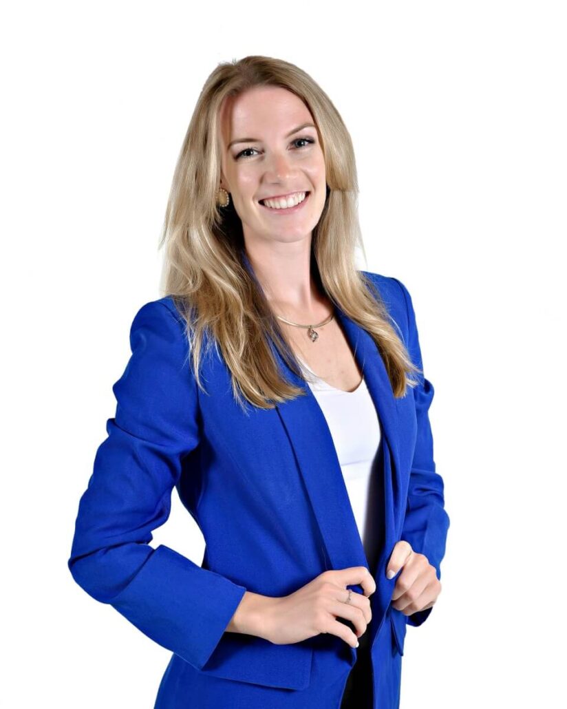 A woman with long blonde hair wearing a bright blue blazer over a white top stands against a plain white background, smiling and facing the camera, ready to take on her 2025 role as Carnival cruise director.