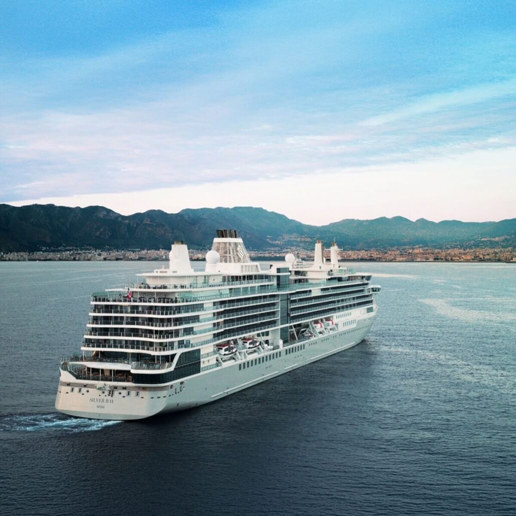 A large white cruise line ship sails near the coastline with mountains in the background under a partly cloudy sky. The ship has multiple decks and lifeboats visible along the side.