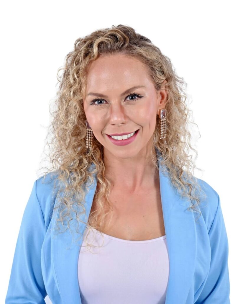 A woman with curly blonde hair wearing a light blue blazer over a white top, smiling at the camera. She has pearl earrings and is posed against a plain white background, reminiscent of a Carnival Cruise Director Schedule for 2025 on a cruise ship.