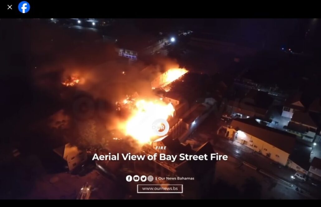 Aerial night view of the Nassau Blaze on Bay Street, with bright orange flames engulfing multiple downtown buildings near the Cruise Port and smoke rising above nearby structures. Text on image reads "Aerial View of Bay Street Fire.