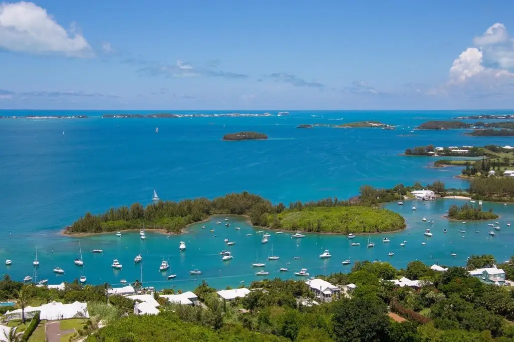 Aerial view of a tropical bay in Bermuda with clear blue water, small green islands, anchored sailboats, and white buildings surrounded by lush vegetation under a partly cloudy sky—a dream escape for any cruise guest.