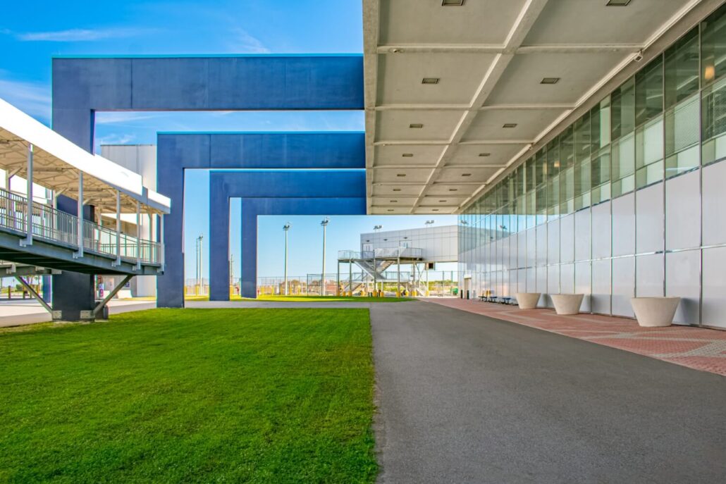 A modern building with large blue geometric structures at Port Canaveral features a glass facade, pathway, green lawn, and metal pedestrian bridge—showcasing cruise terminal upgrades under a clear blue sky.