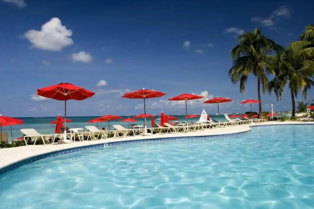 A swimming pool with clear blue water is surrounded by white lounge chairs and red umbrellas. Palm trees and a sandy beach evoke the beauty of the Cayman Islands, with the ocean, blue sky, and cruise dock tenders visible in the distance.