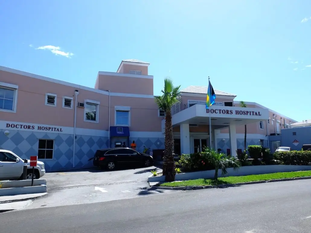 A pink and blue building with a sign reading "Doctors Hospital" above the entrance, where a cruise passenger in critical condition arrived, several cars parked in front, and a flag flying by the main entrance on a sunny day.