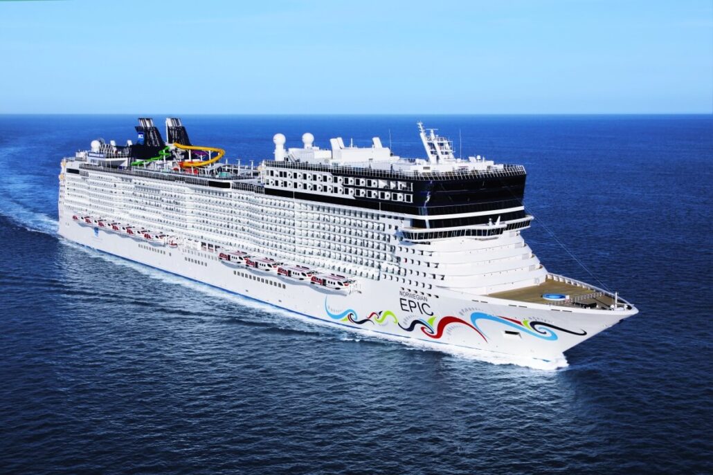 A large white cruise ship labeled "EPIC" sails on calm blue ocean water under a clear sky. The Norwegian Epic features multiple decks, outdoor pools, colorful designs near the bow, new suites, and exclusive adults-only spaces.