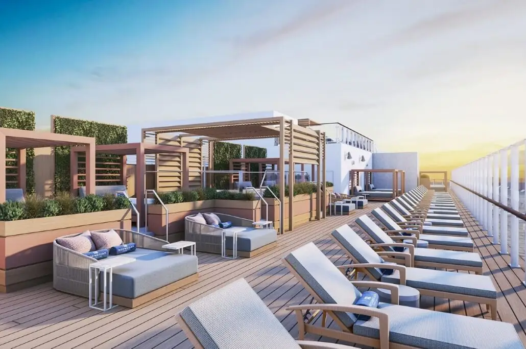 A rooftop deck on the Norwegian Epic features lounge chairs and shaded cabanas, wooden flooring, glass railings, greenery, and towels on each chair—an inviting adults-only space to unwind under a clear sunset sky.