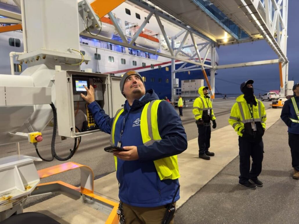 A person in a yellow safety vest operates a control panel at Port Canaveral, with several others nearby. A large ship and overhead structure in the background highlight ongoing cruise terminal upgrades.