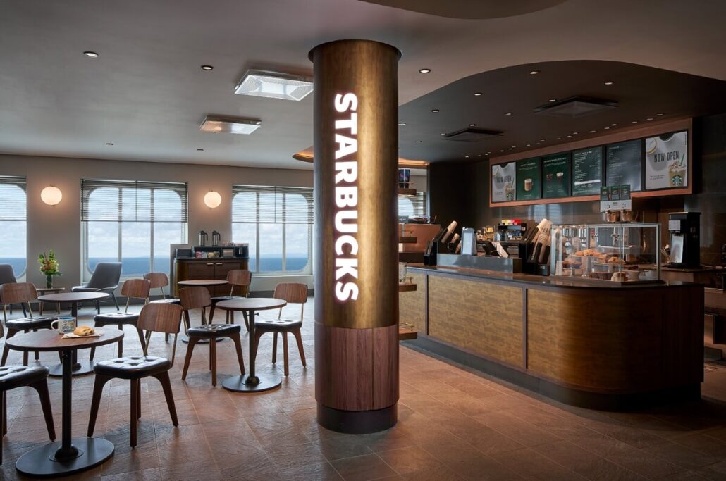 Interior of a Starbucks café with wooden tables and chairs, a central column displaying the Starbucks logo, and menu boards on the wall—reminiscent of the inviting adults-only spaces found aboard Norwegian Epic. Large windows let in natural light.