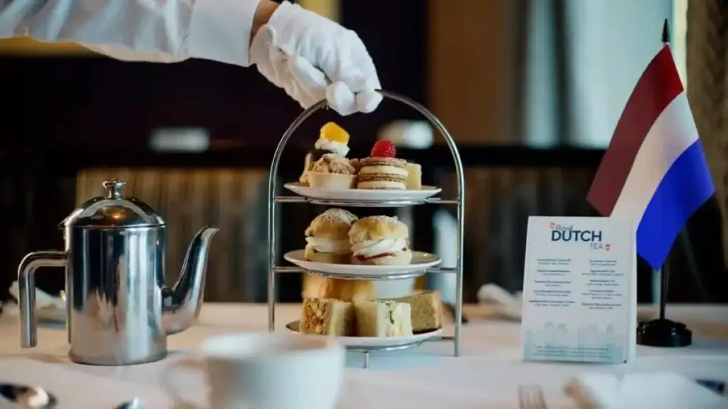 A gloved hand places pastries on a two-tiered tray for a Royal Dutch Tea, with assorted desserts and sandwiches beside a metal teapot, menu card, and a small Dutch flag—perfect for Sea Days aboard Holland America.