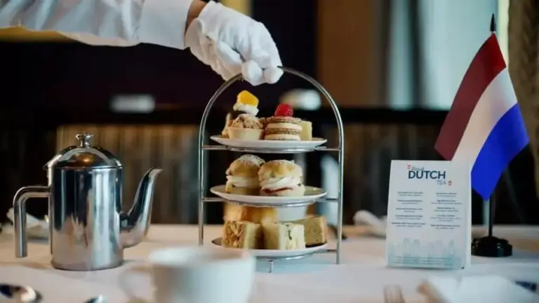 A gloved hand places pastries on a two-tiered tray for a Royal Dutch Tea, with assorted desserts and sandwiches beside a metal teapot, menu card, and a small Dutch flag—perfect for Sea Days aboard Holland America.