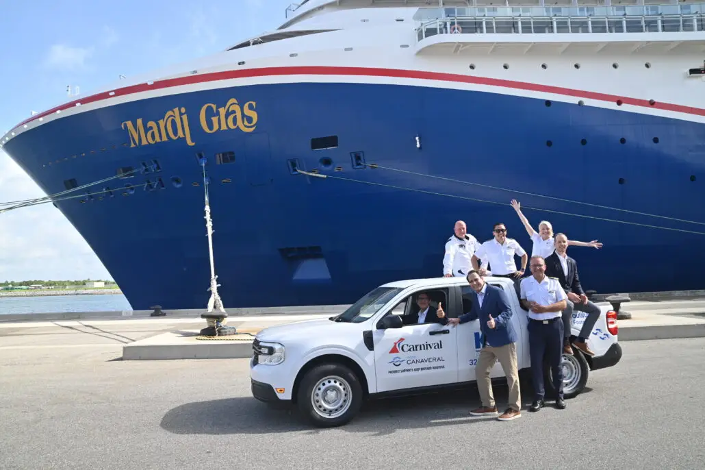 A group of people stands and sits in and around a white Carnival-branded truck parked in front of the large cruise ship Mardi Gras at a dock. Some individuals are waving and smiling toward the camera.