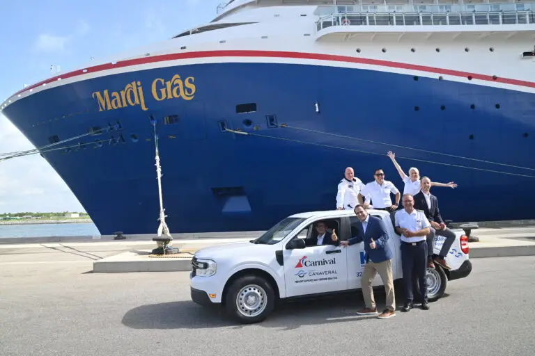 A group of people stands and sits in and around a white Carnival-branded truck parked in front of the large cruise ship Mardi Gras at a dock. Some individuals are waving and smiling toward the camera.