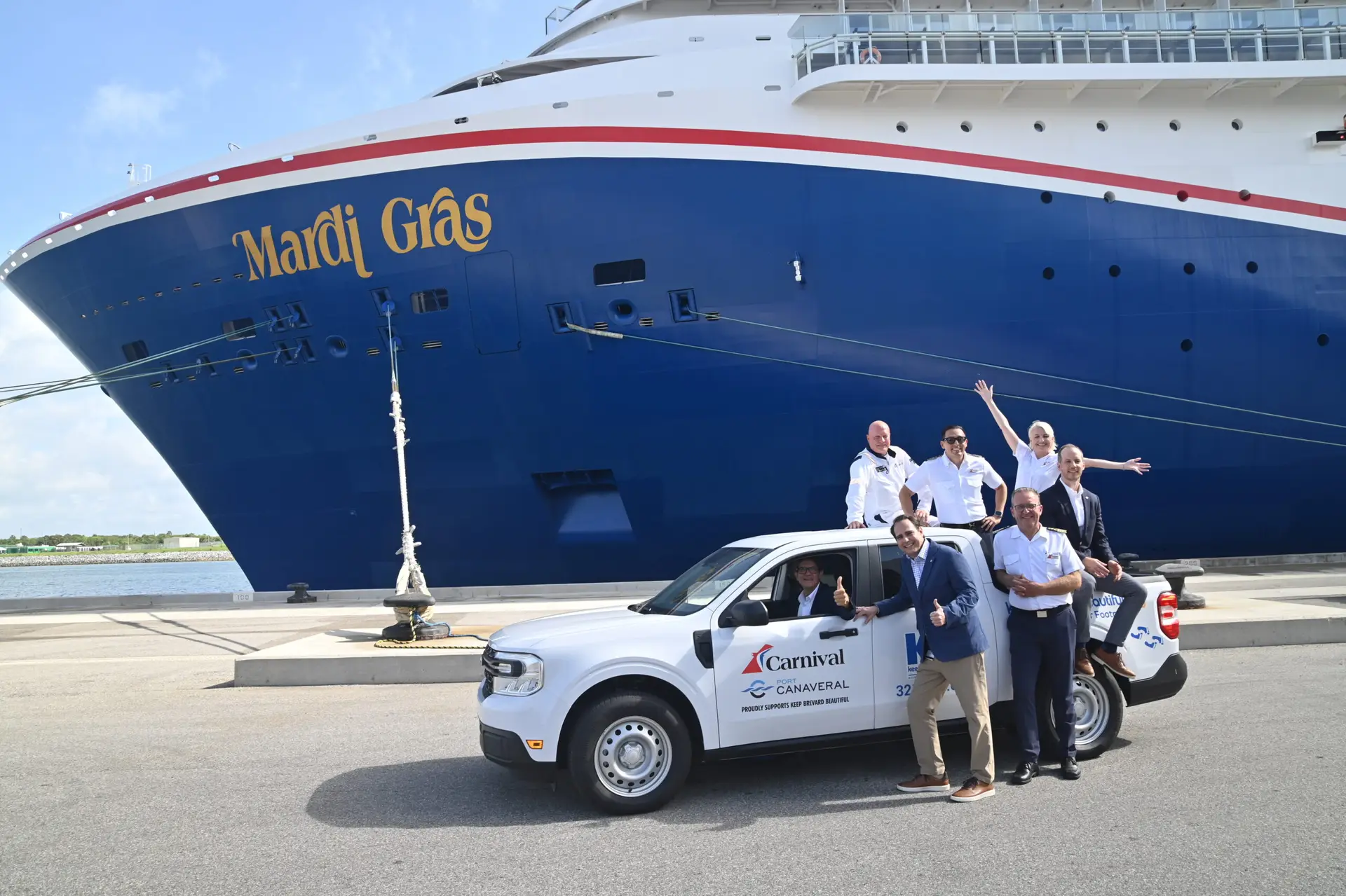A group of people stands and sits in and around a white Carnival-branded truck parked in front of the large cruise ship Mardi Gras at a dock. Some individuals are waving and smiling toward the camera.