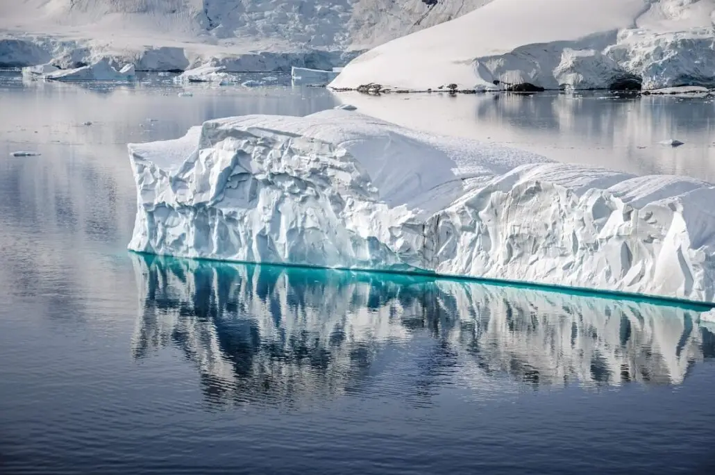 A large, jagged iceberg floats in calm, reflective water with snow-covered land and ice in the background under a clear sky—an awe-inspiring scene typical of an Antarctica Expedition Cruise.