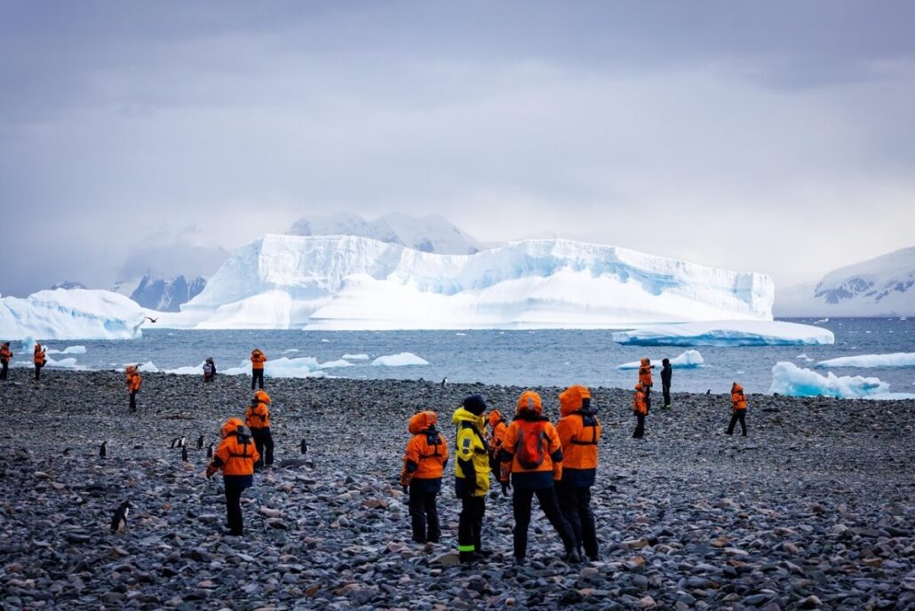 A group of people wearing orange jackets walk on a rocky shore near the ocean during an Antarctica Expedition Cruise, with icebergs and snow-covered mountains in the background under a cloudy sky.