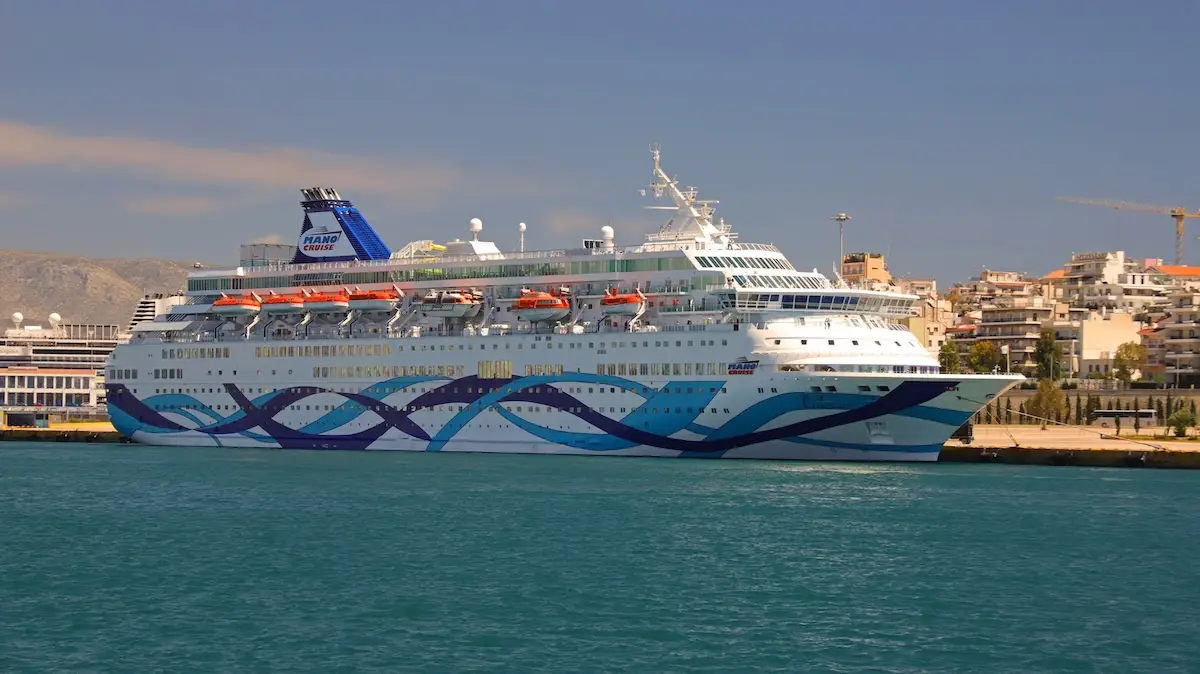 A large white and blue cruise ship with “Panagia Theotokos” written on the funnel is docked in a port, with buildings and cranes visible in the background under a clear sky.